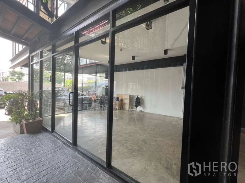 Retail Space for rent - Wide glass façade under canopy with bright interior; potted plants and parked cars outside.