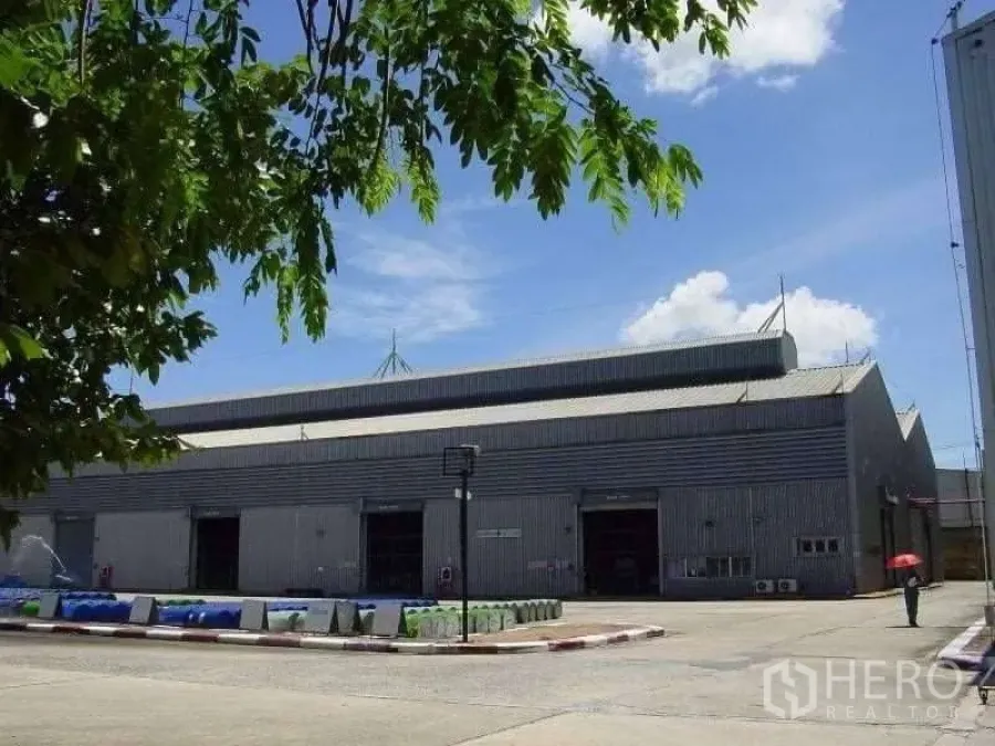 Factory for sale in Rayong - Large metal-clad factory with high gabled roof, multiple roller shutter doors and a wide concrete yard under blue sky, tree framing the foreground.