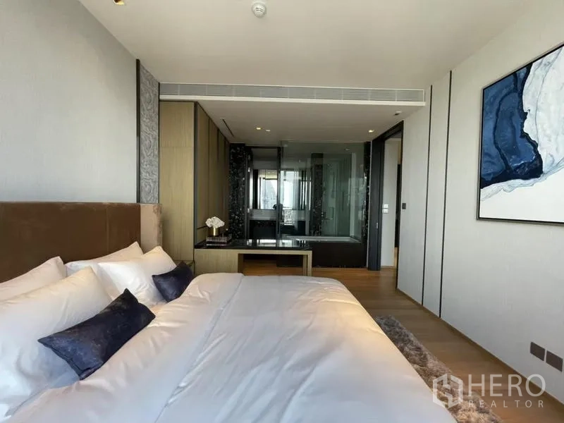 Condominium for rent in Khlong Toei, Bangkok - Primary bedroom facing ensuite bathroom with glass partition and wall art, warm wood flooring throughout.