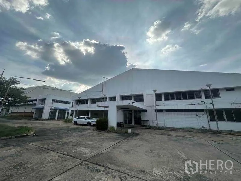 Factory for rent in Khlong Luang, Pathum Thani - Two-story white factory buildings with driveway and parking under a cloudy sky