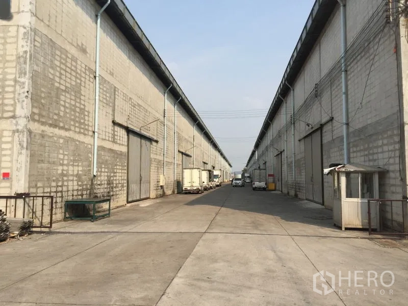 仓库 for rent in Mueang Pathum Thani, Pathum Thani - 两排仓库之间的长通道，可见卡车与小型岗亭。