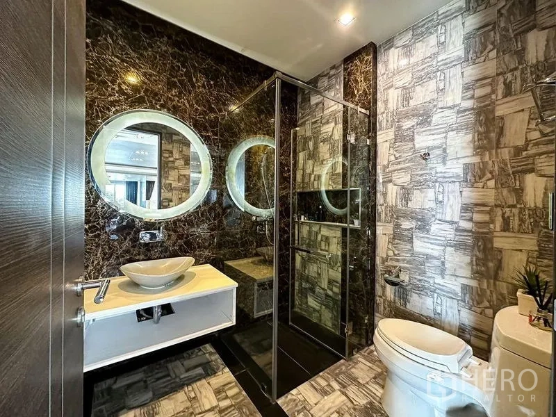Condominium for rent or sale in Khlong Toei, Bangkok - Stylish guest bathroom with glass shower, round illuminated mirrors and patterned tiles.