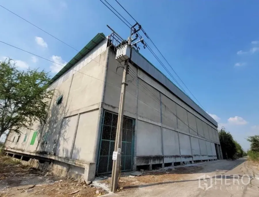 คลังสินค้า ให้เช่าหรือขายในSai Noi, Nonthaburi - มุมอาคารโกดังคอนกรีตหลังคาสีเขียว พร้อมเสาไฟและถนนด้านข้างในไทรน้อย