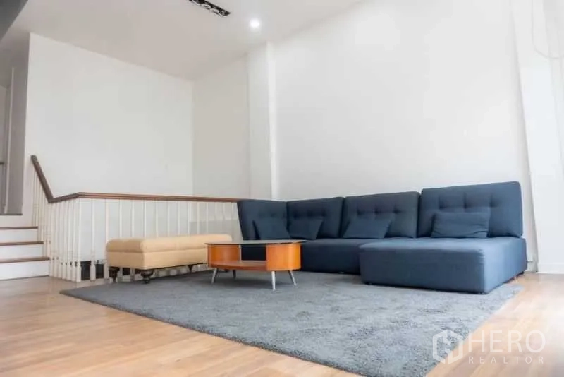 Townhouse for rent in Bang Kapi, Bangkok - Spacious split-level living room with a large L-shaped sofa, gray rug, coffee table, and staircase.