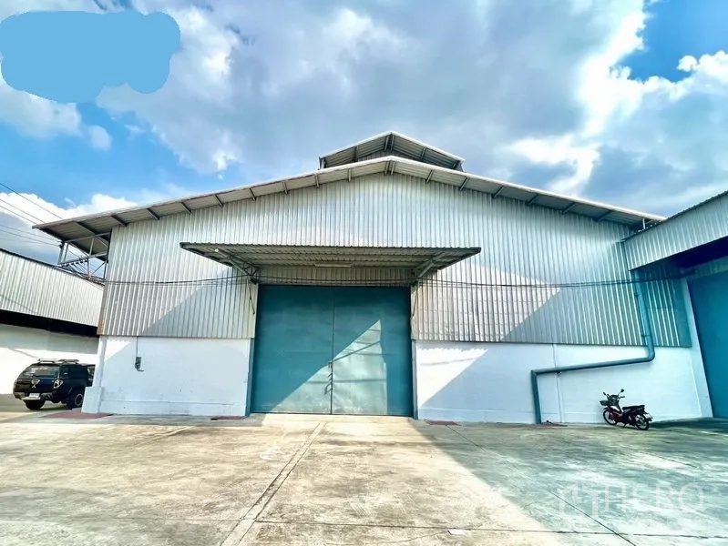 Warehouse for rent in Pak Kret, Nonthaburi - Front exterior of a corrugated-metal warehouse in Pak Kret with a wide concrete yard and large blue sliding door under an awning.