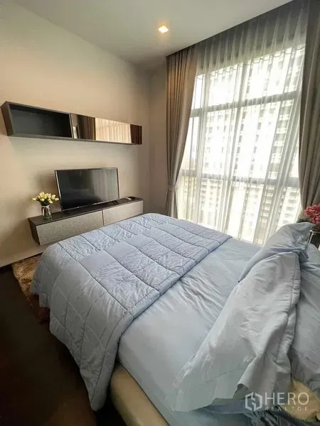 Condominium for rent or sale in Watthana, Bangkok - Bright bedroom with floor-to-ceiling windows, TV console and soft blue bedding with city view beyond sheer curtains.