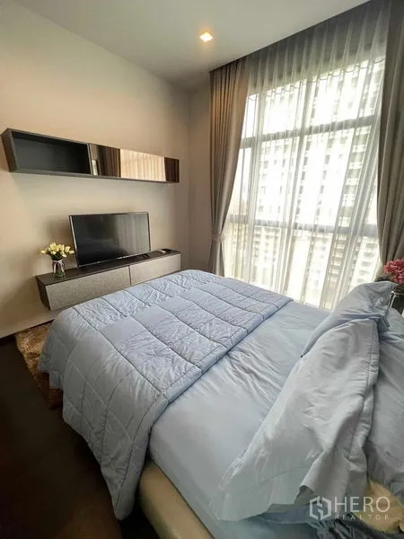 Condominium for rent or sale in Watthana, Bangkok - Bright bedroom with floor-to-ceiling windows, TV console and soft blue bedding with city view beyond sheer curtains.