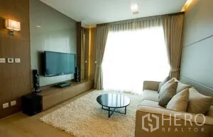 Condominium for rent in Khlong Toei, Bangkok - Bright living room with wall-mounted TV, media console, shag rug, and round coffee table by a large window.