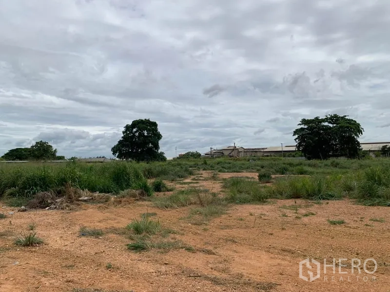 工厂 出租在Chon Buri - 工厂旁大片空地，土质地面与草丛，远处可见建筑。