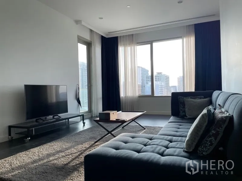 Condominium for rent or sale in Pathum Wan, Bangkok - Living room with sectional sofa, rug and TV stand beside floor-to-ceiling windows facing the skyline.
