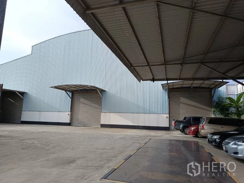 Factory for sale in Ban Phaeo, Samut Sakhon - Corrugated metal factory exterior with wide covered loading bay, roller shutter doors and car parking.