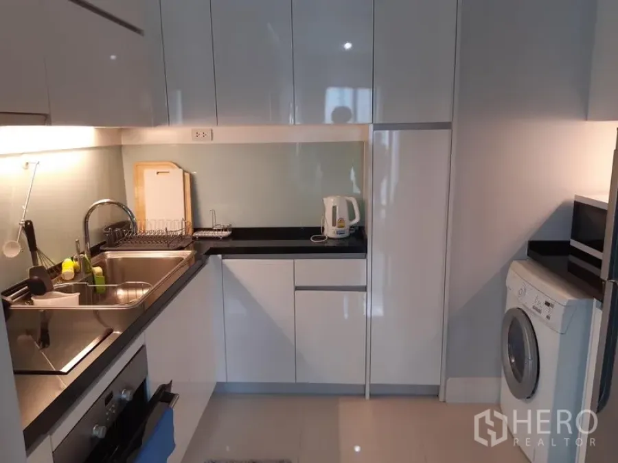 Condominium for rent or sale in Khlong Toei, Bangkok - Modern condo kitchen with glossy white cabinets, black countertops, sink, oven and washing machine.