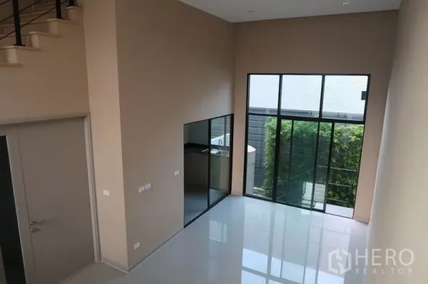Townhouse for rent in Suan Luang, Bangkok - Bright living room with tall windows to a small garden and a glass-partitioned kitchen.