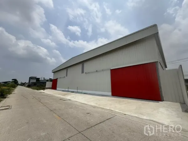 货栈 出租在Lam Luk Ka, Pathum Thani - 仓库侧立面，长条外墙、红色大门与宽阔混凝土车道，方便货车出入。