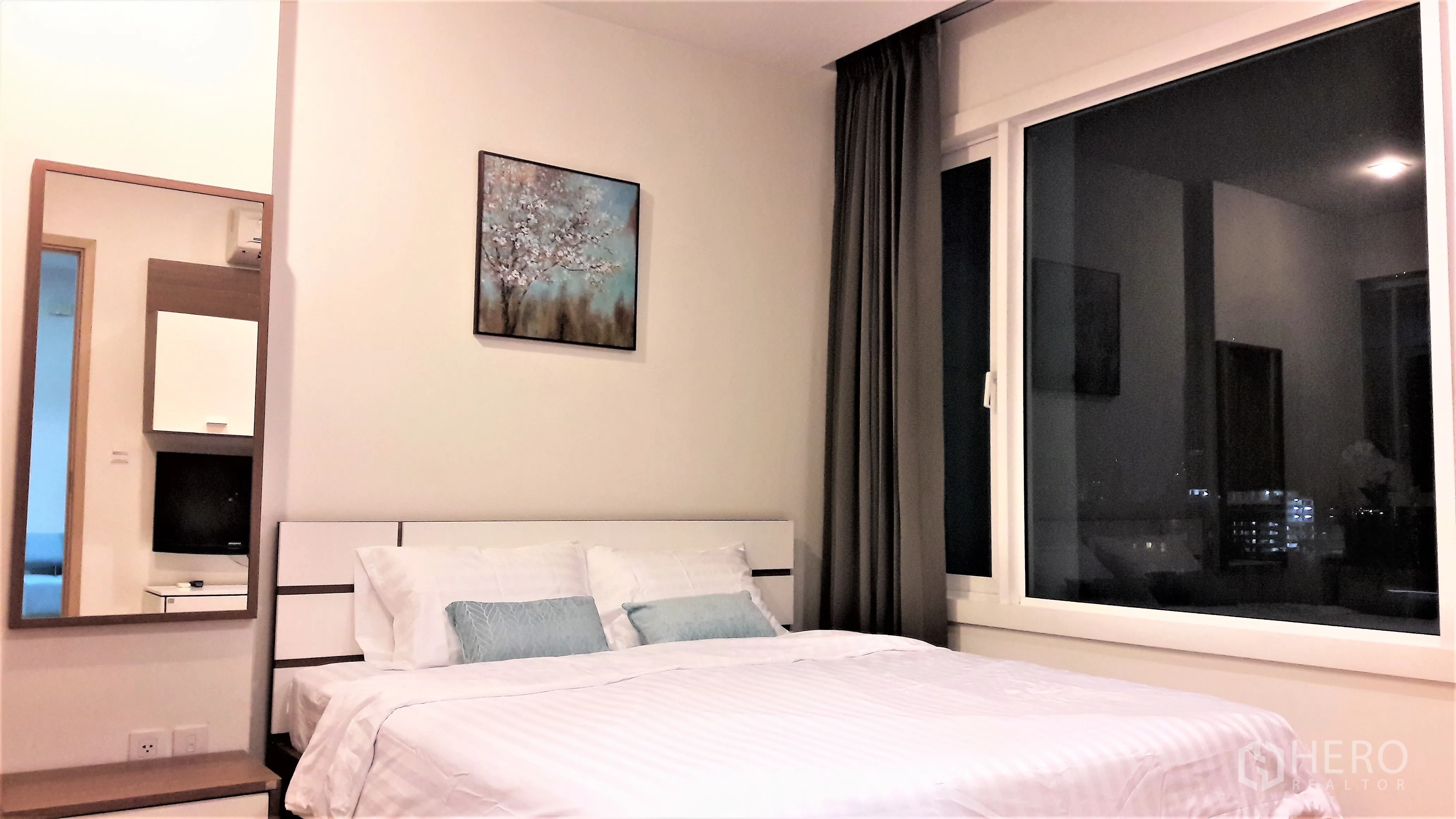 Condominium for rent in Khlong Toei, Bangkok - Bright bedroom with large window, city night view, artwork above a white bed and blackout curtains.