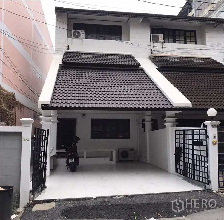 Townhouse for rent in Watthana, Bangkok - Two-storey white townhouse exterior with dark tiled roof, gated entry and covered carport.