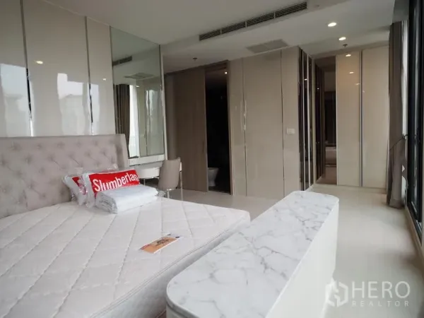 Condominium for rent in Pathum Wan, Bangkok - Bedroom with upholstered bed, marble-top bench, vanity and doorway to bathroom.