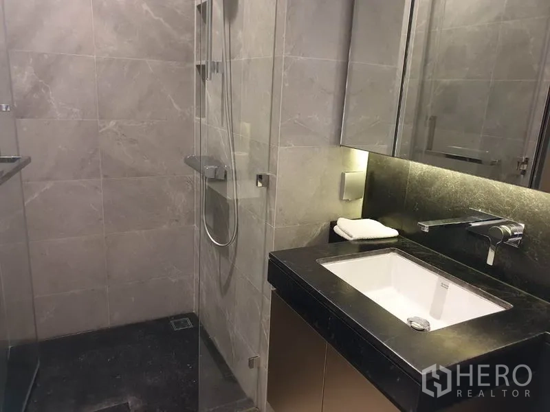 Condominium for rent in Pathum Wan, Bangkok - Modern bathroom with glass-enclosed shower, stone tiles and underlit vanity.