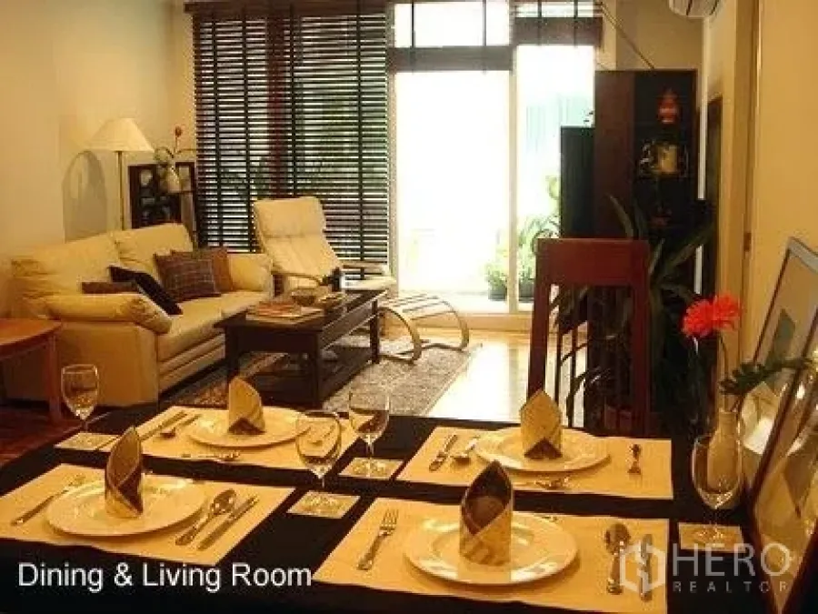 Condominium for rent in Khlong Toei, Bangkok - Warm dining and living room with set table, sofa and media cabinet, opening to a balcony via sliding glass doors.
