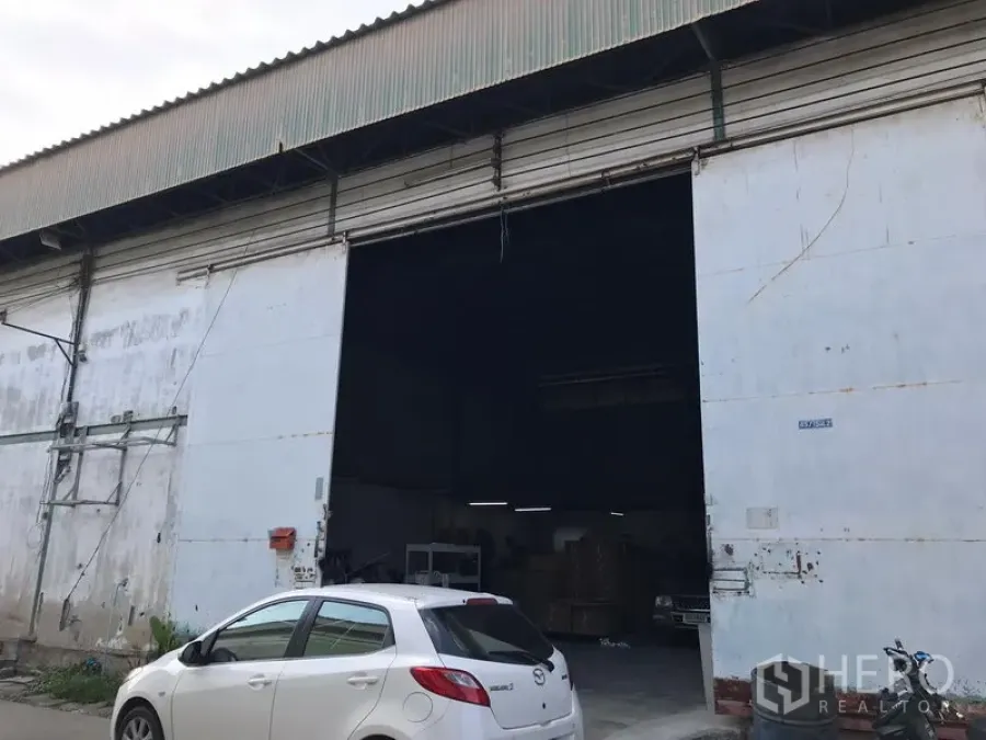 货栈 出租在Bang Phli, Samut Prakan - 仓库外立面，超宽金属卷闸门，高挑入口，并设有前方停车与装卸区。