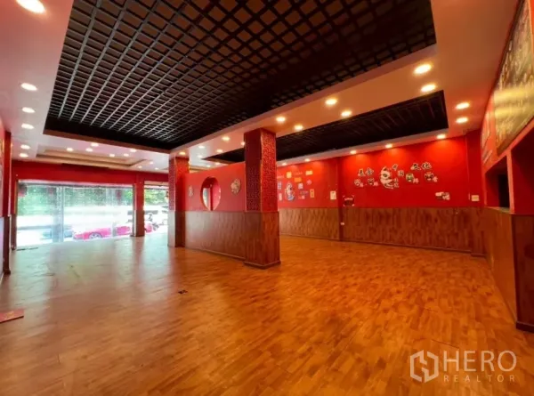 Shophouse for rent in Watthana, Bangkok - Spacious ground-floor retail hall with wood floors, red walls, and a black grid ceiling with lights.