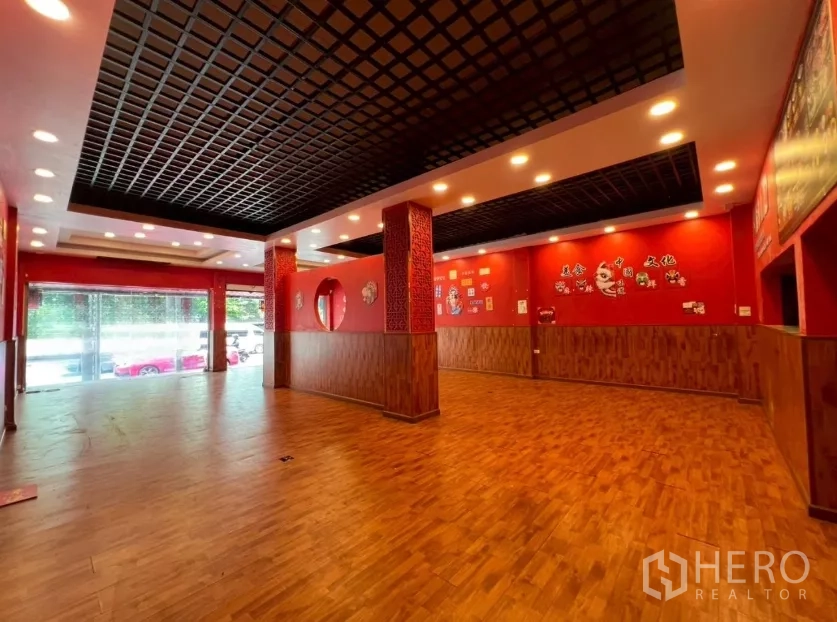 Shophouse for rent in Watthana, Bangkok - Spacious ground-floor retail hall with wood floors, red walls, and a black grid ceiling with lights.