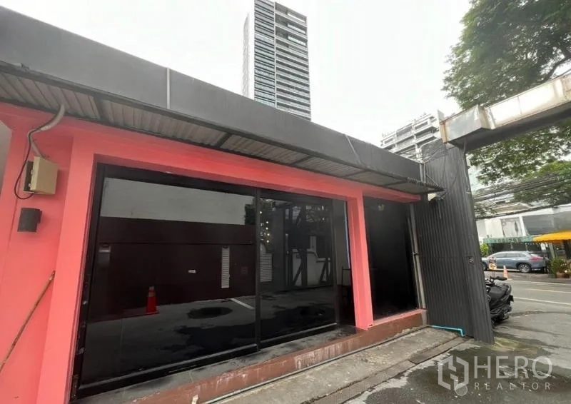 Retail Space for rent in Watthana, Bangkok - Single-story retail facade with wide black glass window framed in pink, with high-rise in background.