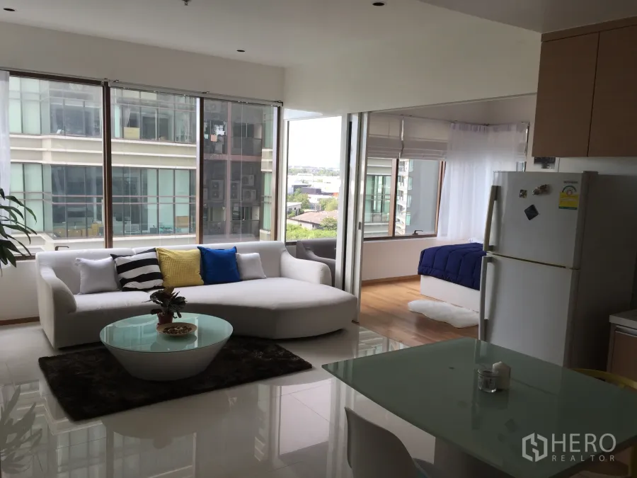 Condominium for rent in Khlong Toei, Bangkok - Bright open‑plan living area with white sofa, glass coffee table, dining corner and large windows with city views.