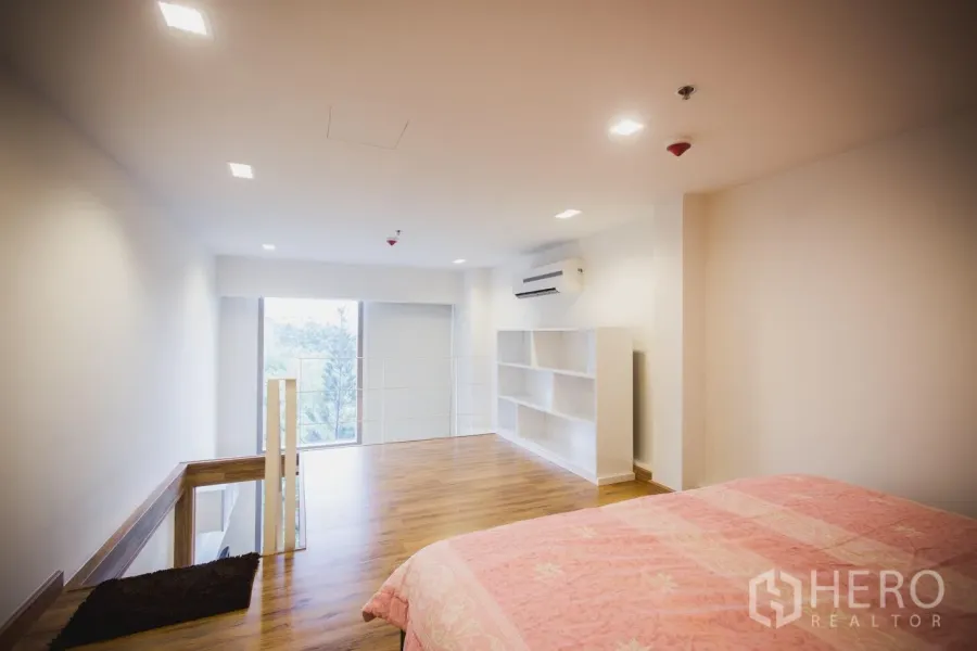 Condominium for rent in Khlong Toei, Bangkok - Bright loft-style bedroom with mezzanine, wood floors, built-in shelving, air conditioner and large window with leafy view.