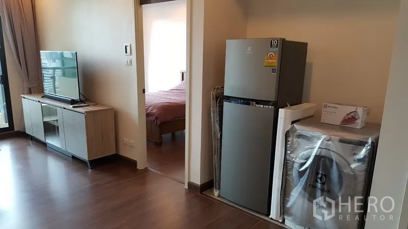 Condominium for rent in Sathon, Bangkok - Living area with TV cabinet, stainless refrigerator and washing machine by bedroom doorway.