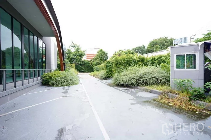 Standalone for rent in Bang Na, Bangkok - Side exterior walkway beside a glass curtain wall and landscaped hedges.