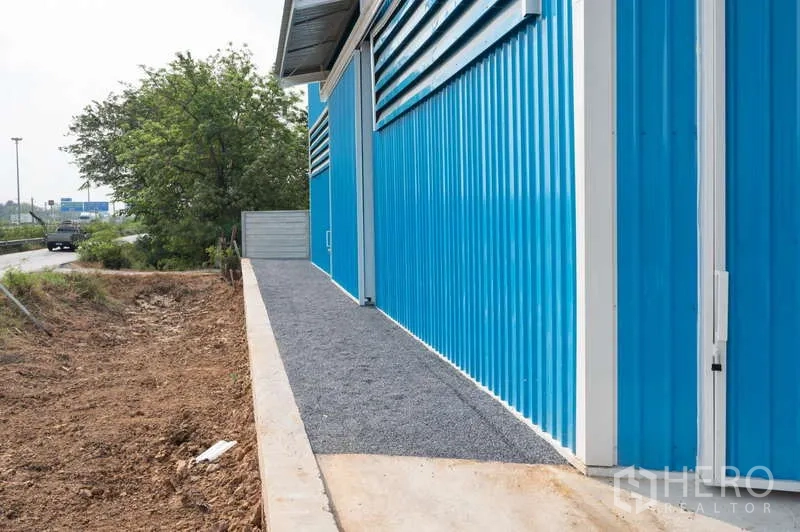 Warehouse for rent in Khlong Luang, Pathum Thani - Side elevation of the blue warehouse with a gravel walkway beside a roadside verge and trees.