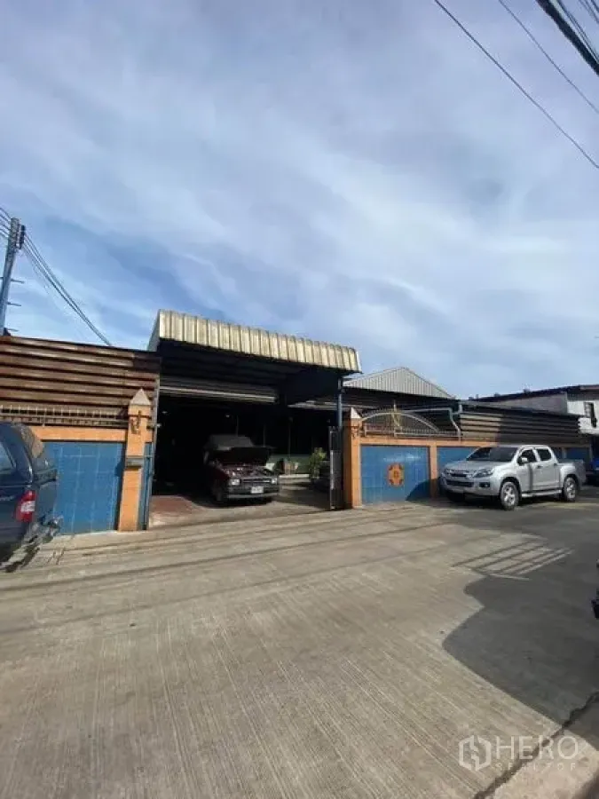 Factory for sale in Bang Khun Thian, Bangkok - Street-facing factory entrance with blue tiled wall, rolling gate and covered parking for pickup trucks.