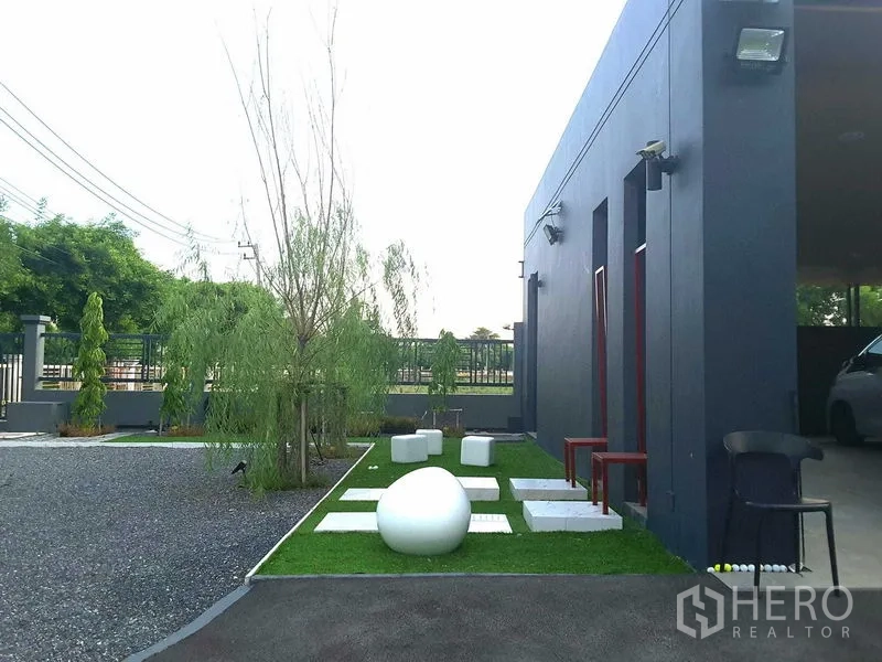 Detached House for rent or sale in Lam Luk Ka, Pathum Thani - Side yard with stepping stones on green lawn, sculptural seats and slender trees beside a grey wall.