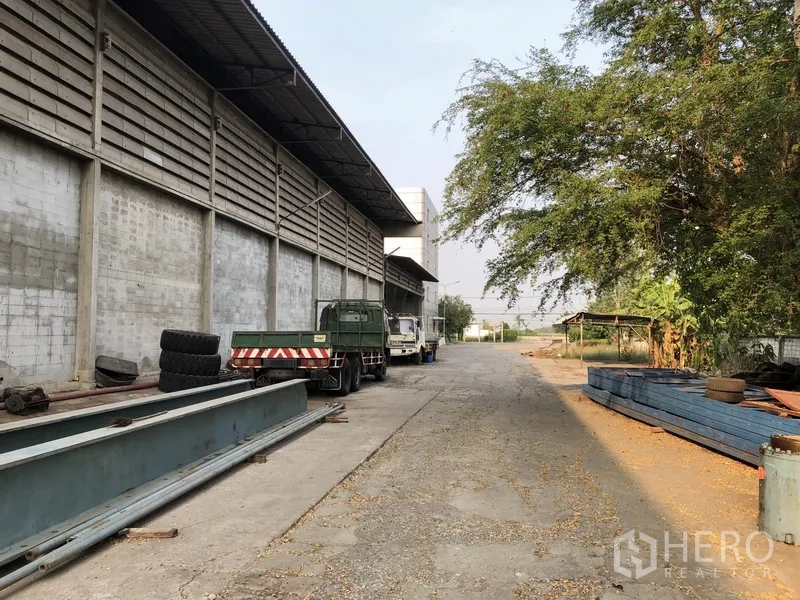 โรงงาน ให้เช่าในBang Bua Thong, Nonthaburi - ลานด้านข้างอาคารโรงงาน มีรถบรรทุก คานเหล็ก และต้นไม้ใหญ่
