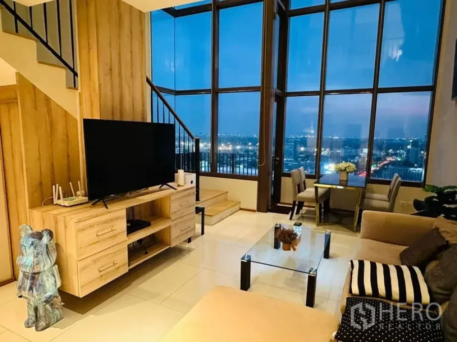 Condominium for rent in Bangkok - Living room with TV unit, glass coffee table and panoramic window wall at night.