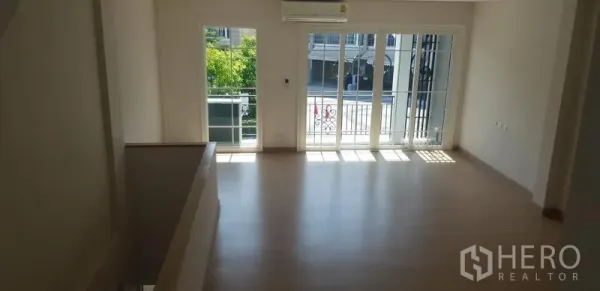 Townhouse for rent in Bang Na, Bangkok - Sunlit upper floor room with light wood flooring and sliding glass doors opening to a balcony.