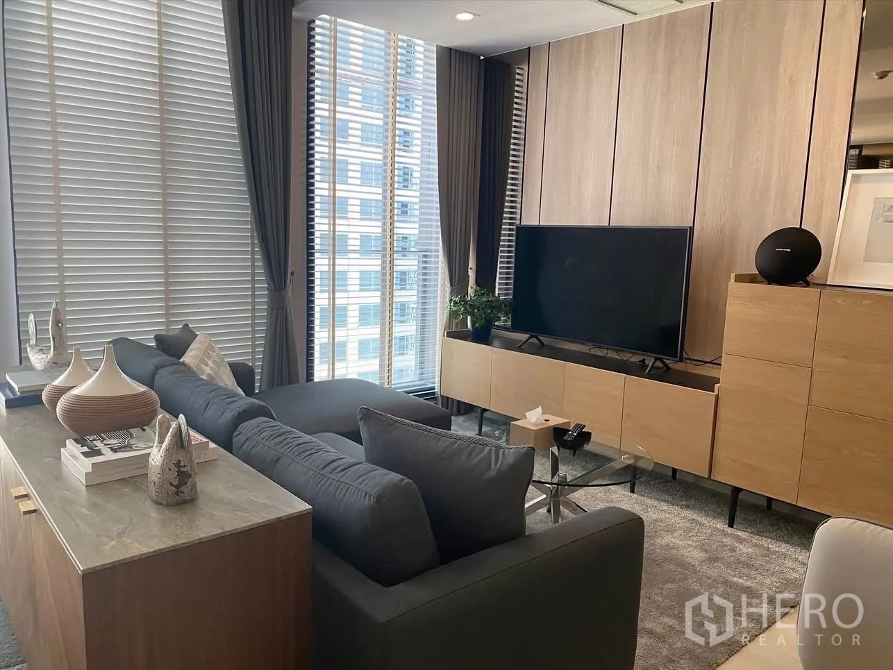 Condominium for rent in Pathum Wan, Bangkok - Living room with gray sectional sofa, TV console, and tall windows with blinds.