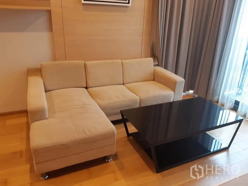 Condominium for rent in Khlong Toei, Bangkok - Beige L-shaped sofa beside a large window with a sleek black glass coffee table in the living room.