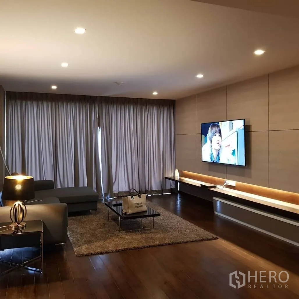 Condominium for rent or sale in Pathum Wan, Bangkok - Cozy living room with sectional sofa, wall-mounted TV, recessed lighting, and full-length curtains.