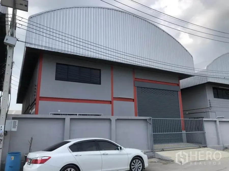 Warehouse for rent in Nonthaburi - Modern grey-and-red warehouse exterior with curved metal roof, large roller shutter and gated entrance on a concrete road in an industrial area.