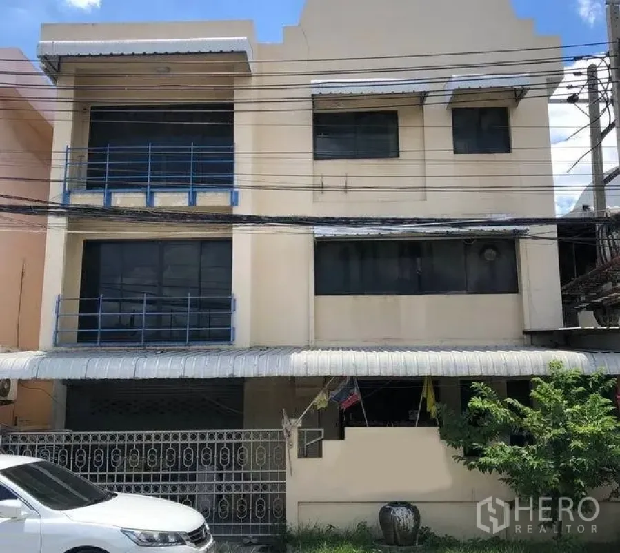 Factory for sale in Bang Bua Thong, Nonthaburi - Three-storey beige factory facade with blue railings, gated parking and a covered frontage under power lines in Bang Bua Thong, Nonthaburi.