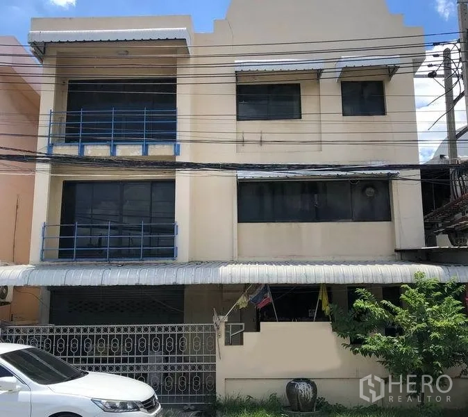 Factory for sale in Bang Bua Thong, Nonthaburi - Three-storey beige factory facade with blue railings, gated parking and a covered frontage under power lines in Bang Bua Thong, Nonthaburi.