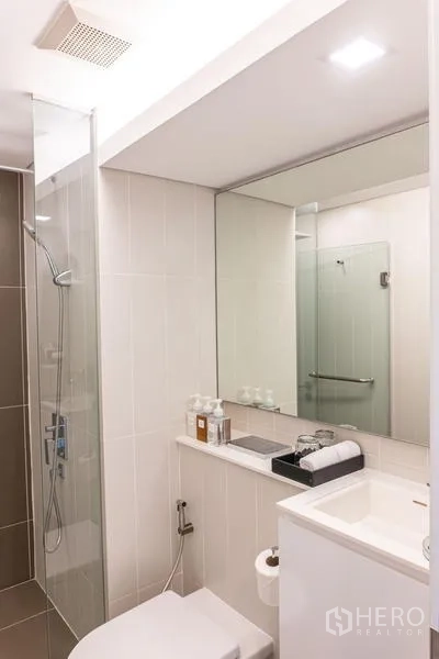 Condominium for rent in Khlong Toei, Bangkok - Clean bathroom with large mirror, vanity, toilet and a glass‑enclosed shower.