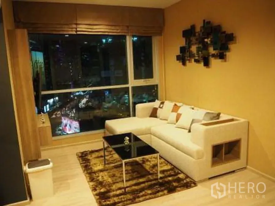 Condominium for rent in Sathon, Bangkok - Sathon condo living room with L-shaped sofa, rug and floor-to-ceiling window showing Bangkok night city view.