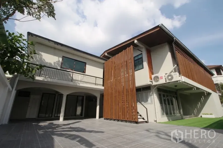Detached House for rent in Chatuchak, Bangkok - Rear courtyard view of two‑storey home with vertical timber screens, balcony, tiled patio, and small lawn under blue sky.