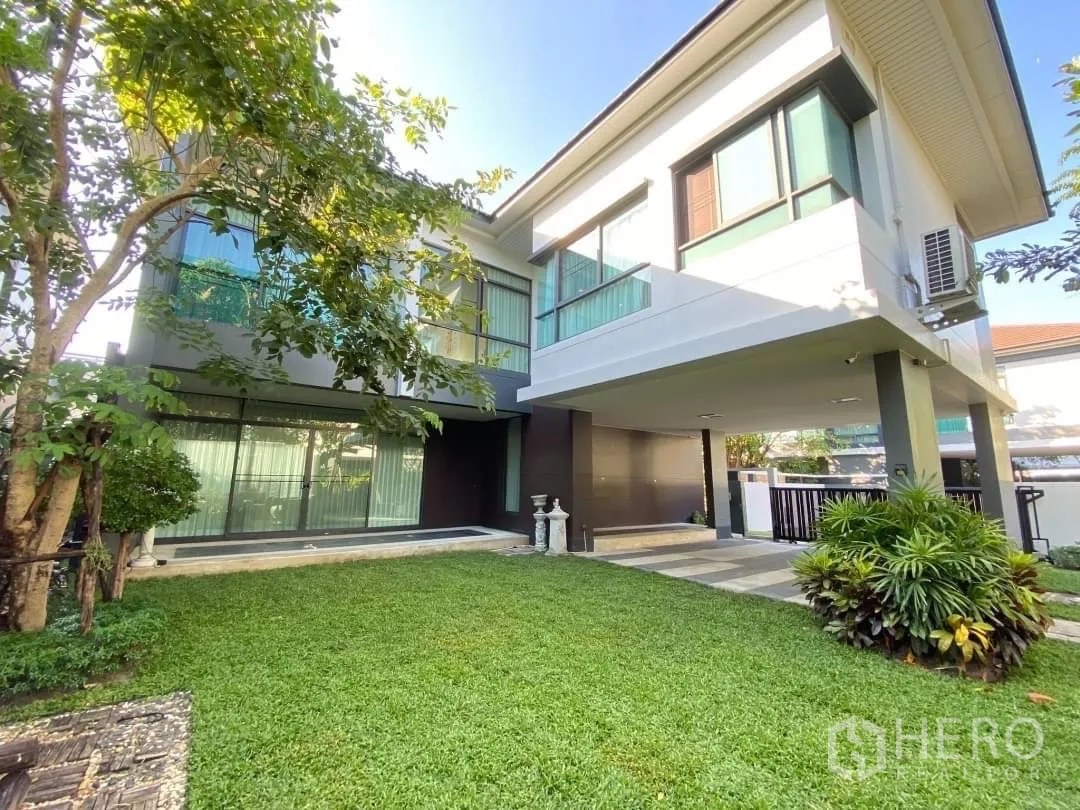 Detached House for rent or sale in Bang Kapi, Bangkok - Side view of the home with covered carport, large windows and a lush green lawn with trees.