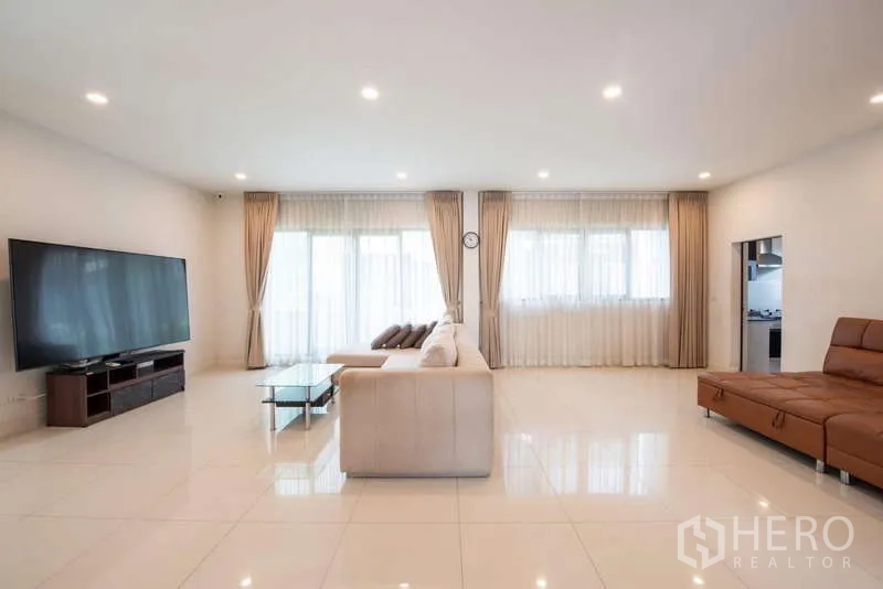 Detached House for rent or sale in Kanchanaburi - Open-plan living room with large TV, sectional sofa, and wide windows with sheer curtains.