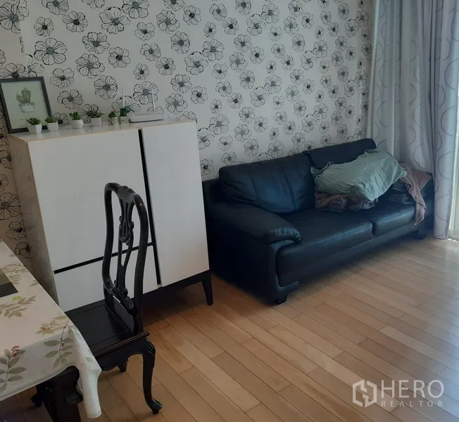 Condominium for rent in Khlong Toei, Bangkok - Cozy living area with black leather sofa, floral wallpaper, low cabinet and light wood flooring beside balcony curtains.