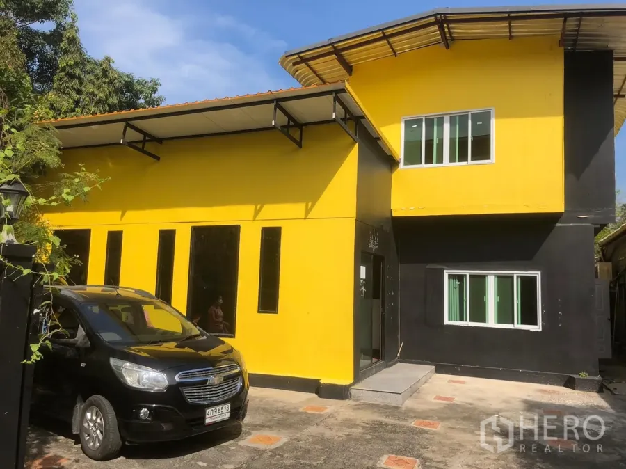 独立式住宅 出租在Khlong Toei, Bangkok - 黄黑配色的两层独栋住宅，滑动窗户，院内铺装车道与停车位，天空晴朗。